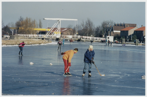 Eine Gruppe von Menschen, die Eis hockey auf einem zugefrorenen See spielt, umgeben von Bäumen, Gebäuden, Polen und einer Brücke, mit Straßenlaternen, die die Szene beleuchten und der Himmel oben sichtbar ist.