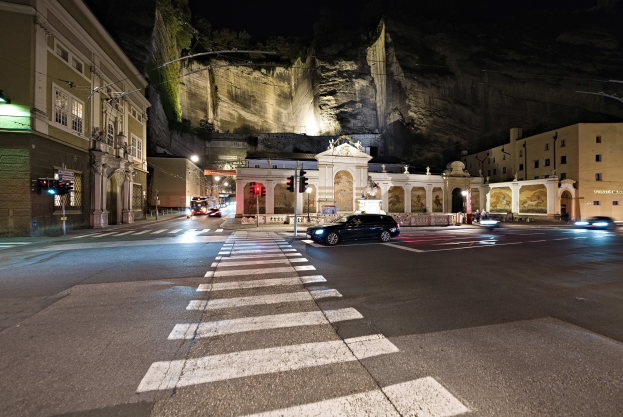 Eine nächtliche Stadtstraße mit fahrenden Autos, Straßenlaternen, Ampeln, Gebäuden mit beleuchteten Fenstern, Laternenmasten, einem Hügel im Hintergrund und einer Zebrastreifenkreuzung unten.