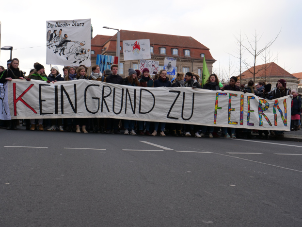 Eine Gruppe von Menschen mit einem Transparent mit der Aufschrift "Kein Grund zu Feiern" demonstriert auf einer Straße mit Laternenmasten, Straßenlaternen, Bäumen, Gebäuden und einem klaren Himmel.
