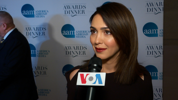Emmy Rossum in einem schwarzen Kleid mit Mikrofon, mit einem Mann zu ihrer Linken und einem Banner mit der Aufschrift "Aam Awards Dinner" im Hintergrund.