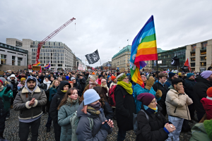 Eine große Gruppe von Menschen bei einer LGBTQ+-Rechtsdemo in Berlin, die Fahnen und Schilder halten, mit Gebäuden, einem Kran und einem bewölkten Himmel im Hintergrund.