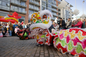 Ein lebendiges chinesisches Neujahrsfest in Amsterdam mit einem Löwen-Tanz im Vordergrund und einer Menge Menschen drumherum, einige halten Kameras, vor einem Hintergrund aus Gebäuden, Laternenmasten und einem klaren blauen Himmel.