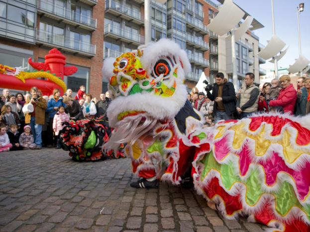 Ein lebendiges chinesisches Neujahrsfest in Amsterdam mit einem Löwen-Tanz im Vordergrund und einer Menge Menschen drumherum, einige halten Kameras, vor einem Hintergrund aus Gebäuden, Laternenmasten und einem klaren blauen Himmel.