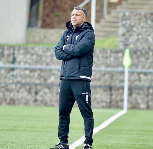 Ein Fussballtrainer mit verschränkten Armen auf einem Feld, der eine schwarze Jacke und Hose trägt, vor einem Zaun, Treppe und Wand.