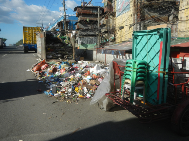Ein Lastwagen steht neben einem Haufen Müll auf einer Straße, mit einem Wagen mit Plastikstühlen rechts daneben und Gebäuden, Strommasten, Bäumen und einem bewölkten Himmel im Hintergrund.