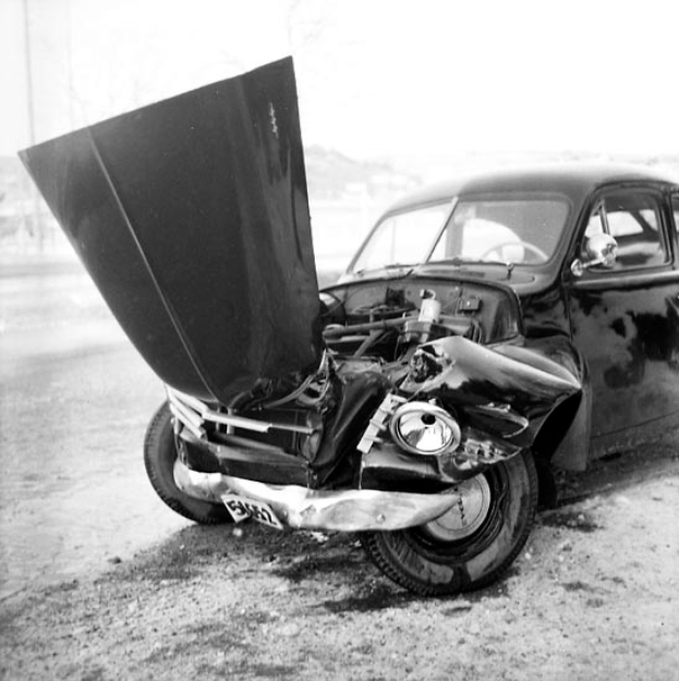 Ein Schwarz-Weiß-Foto eines schwer beschädigten Autos am Straßenrand, mit eingedellter Motorhaube, zersplitterten Fenstern und einer offenen Fahrertür, die den Motor zeigt.
