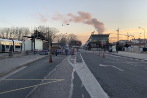 Eine Stadtstraße mit Absperrbaken, einem Bus, Laternen, Bäumen, Gebäuden, Kränen und Rauch, der aus einem entfernten Gebäude aufsteigt, vor einem Himmel.