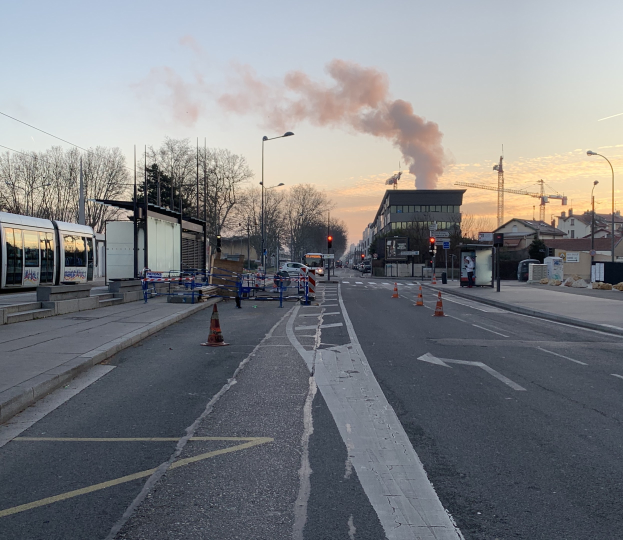 Eine Stadtstraße mit Absperrbaken, einem Bus, Laternen, Bäumen, Gebäuden, Kränen und Rauch, der aus einem entfernten Gebäude aufsteigt, vor einem Himmel.