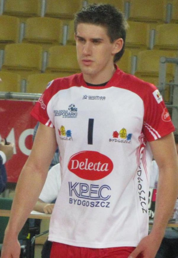Ein Mann in einem roten und weißen Handballtrikot steht auf einem Feld, umgeben von Zuschauern, mit einem roten Banner im Hintergrund, das "Krasnoyarsk vs Bydgoszcz" trägt und entschlossen aussieht.
