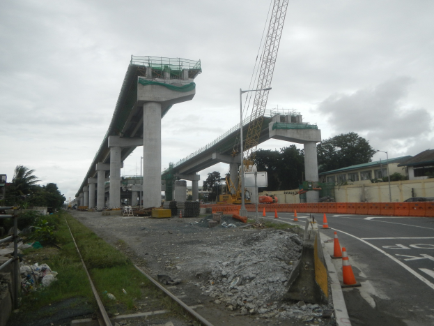 Baustelle mit einer Brücke im Hintergrund, Straße mit Absperrbaken markiert, Bahnschienen auf der linken Seite, verstreute Steine und Gras, Bäume und Gebäude säumen die Straße und ein bewölkter Himmel.