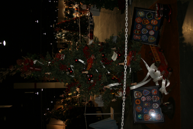 Ein Weihnachtsbaum mit Geschenken, Rahmen und Spielzeugen auf einer Plattform, mit Menschen, zusätzlichen Weihnachtsbäumen, Ketten, Pfosten und Lichtern im Hintergrund.