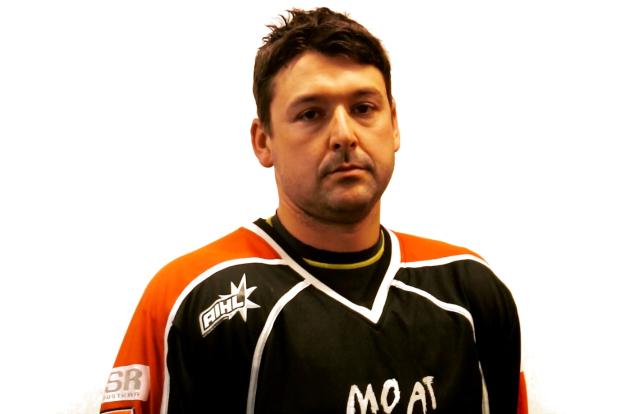Ein Mann in einem schwarzen und orangen Hockey-Jersey mit ernstem Gesichtsausdruck, gefalteten Händen, kurzem dunklen Haar und Bartstoppeln steht vor einem weißen Hintergrund.