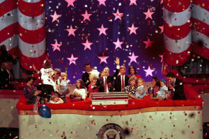 Eine Gruppe von Menschen auf einer Bühne mit einem Rednerpult, Luftballons und Konfetti, mit Sternen und einem Vorhang im Hintergrund.
