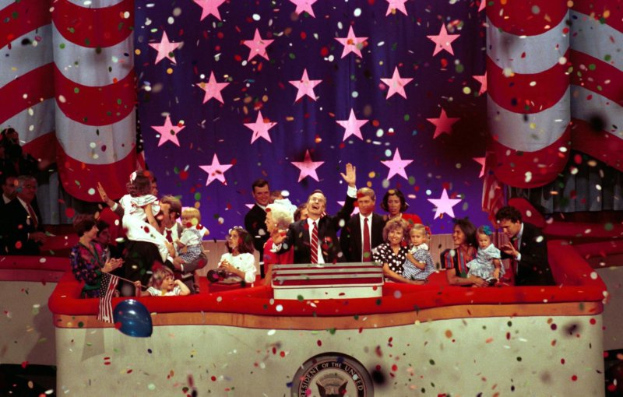 Eine Gruppe von Menschen auf einer Bühne mit einem Rednerpult, Luftballons und Konfetti, mit Sternen und einem Vorhang im Hintergrund.