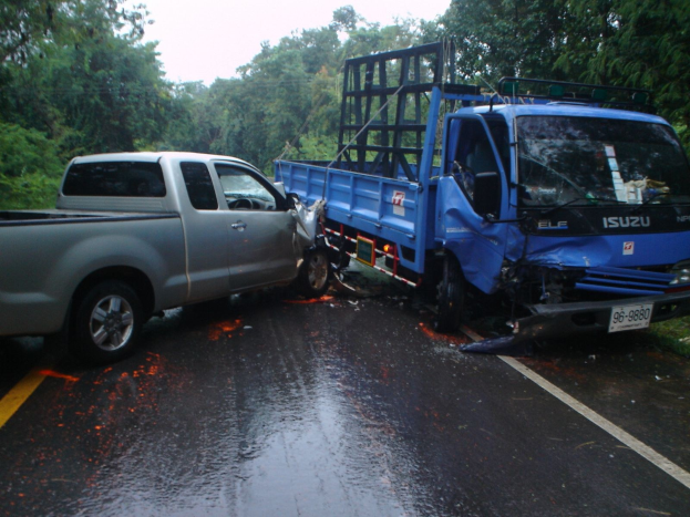 Ein schwerbeschädigter Lkw mit eingedellter Front und verbeulter Karosserie steht neben der Straße, umgeben von Bäumen unter einem klaren blauen Himmel.