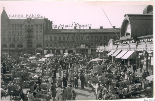 Ein Schwarz-Weiß-Foto von einem belebten Markt in Berlin, Deutschland, mit Menschen, die spazieren gehen, einige mit Schirmen, und Karren mit Gemüse, mit Gebäuden im Hintergrund.