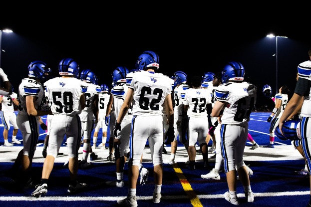 Eine Gruppe von Füßballspielern in Helmen und Stiefeln steht auf einem Feld unter hellen Stadionlichtern und sieht bereit für ein Spiel aus.