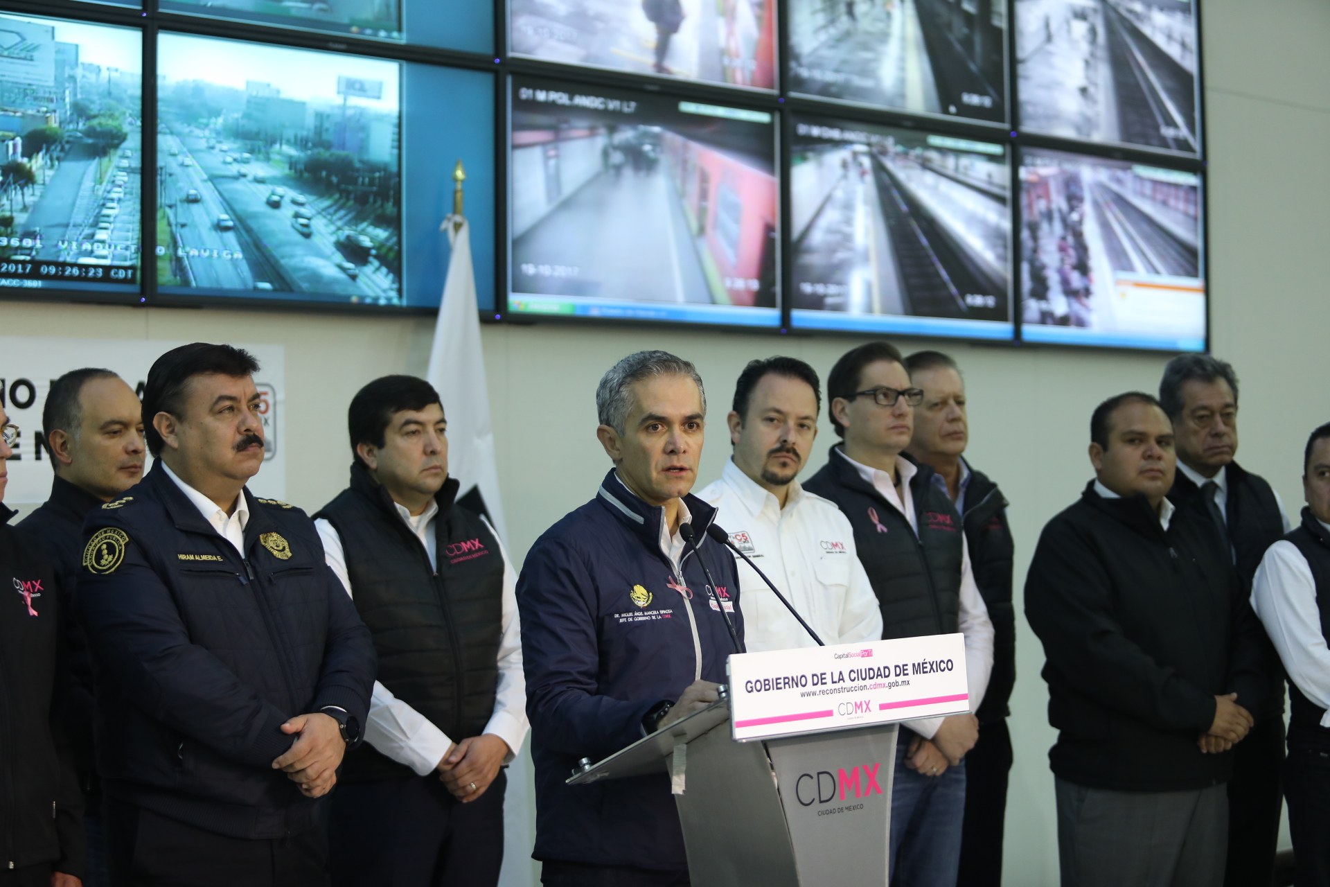 Gruppe von Männern neben einem Rednerpult mit einem Mikrofon und einer Tafel mit der Aufschrift "Mexikanische Zoll- und Grenzschutzbehörde", vor einer Wand mit Fernsehern und einer Flagge.