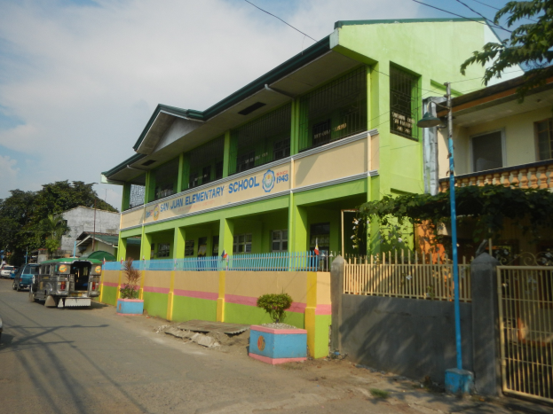 Schulgebäude mit grüner und gelber Fassade, Fahrzeuge auf der Straße, Topfpflanzen, ein Zaun mit einem Tor, ein Straßenpfahl, Bäume, Überlandleitungen und ein bewölkter Himmel.