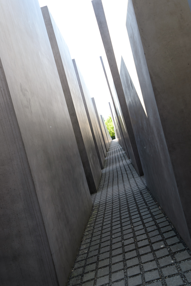 Ein von Betonwänden gesäumter Weg im Holocaust-Mahnmal für die ermordeten Juden Europas in Berlin, Deutschland, mit Bäumen im Hintergrund und einem klaren blauen Himmel darüber.