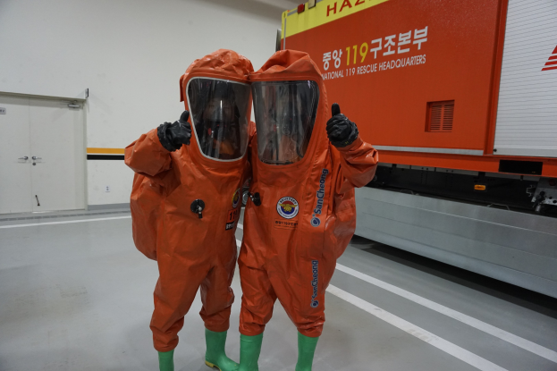 Zwei Personen in orangen Schutzanzügen stehen nebeneinander vor einem Truck, mit einer Wand und Türen im Hintergrund.