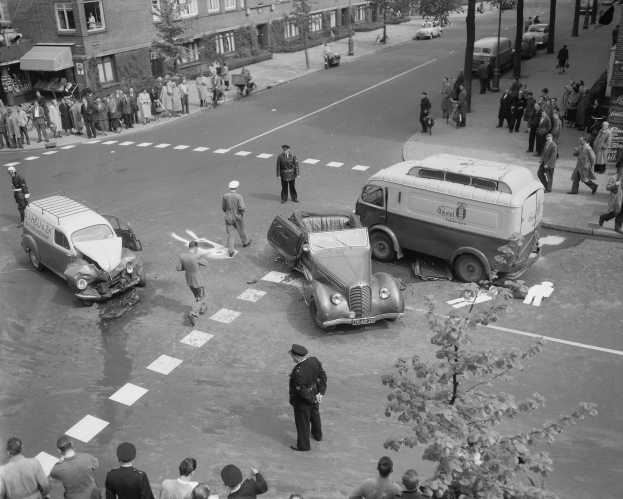 Eine Schwarz-Weiß-Szene eines Autounfalls an einer Straßenecke, mit mehreren Fahrzeugen, darunter ein Van, einer Gruppe von Menschen drumherum und Gebäuden mit Fenstern, Bäumen, Masten und Texttafeln im Hintergrund.