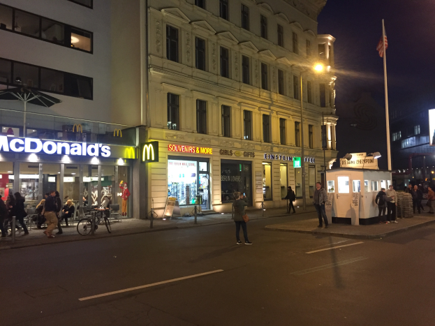Eine Gruppe von Menschen geht nachts eine Straße in Berlin, Deutschland, entlang, gesäumt von Gebäuden, Laternen, einer Flagge, Geschäften mit Schildern, Fahrrädern und anderen Gegenständen, mit dem Himmel im Hintergrund.