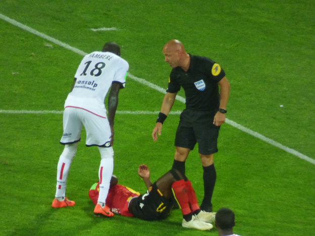 Ein Fußballspieler liegt neben einem Schiedsrichter auf dem Boden, umgeben von drei stehenden Personen auf einem Rasenfeld, wobei eine zentrale Figur mit entschlossener Miene und ausgestreckten Armen zu sehen ist.