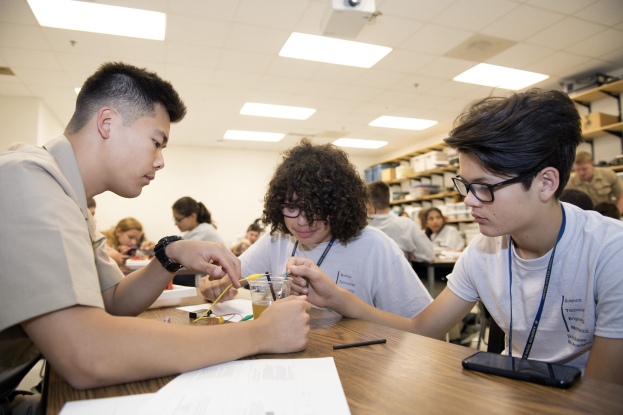 Three students sit at a classroom table working on a project, surrounded by papers, pens, a mobile phone, and other items, with storage racks and ceiling-mounted lights and a projector in the background.