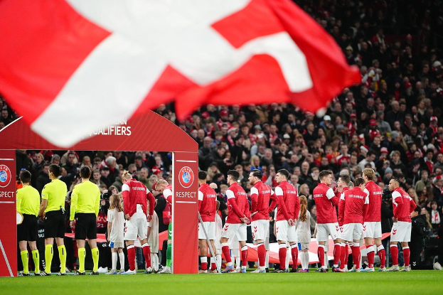 Eine Gruppe von Menschen auf einem Fußballfeld mit einer roten und weißen Fahne im Vordergrund, einem Bogen mit der Aufschrift "Bayern München vs Bayern München Wetten & Vorschau" im Hintergrund und einer großen Menge im Stadion.