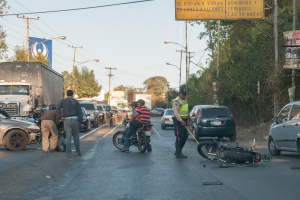Eine Gruppe von Menschen steht um ein verunglücktes Motorrad auf der Straße herum, umgeben von mehreren Fahrzeugen, darunter ein Lastwagen, sowie Bäumen, Pfosten, Laternen und Schildern im Hintergrund unter dem Himmel.