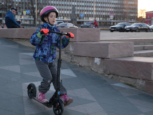 Ein junger Junge fährt mit einem Helm und Handschuhen auf einem Scooter einen Gehweg entlang, mit Stufen, Fahrzeugen, Menschen, Bäumen, Polen, Brettern, Gebäuden und einem klaren blauen Himmel im Hintergrund.