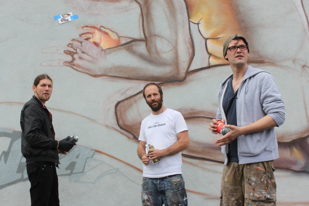 Menschen stehen und halten Farbsprühflaschen in der Hand, mit einer Graffiti-bedeckten Wand im Hintergrund.