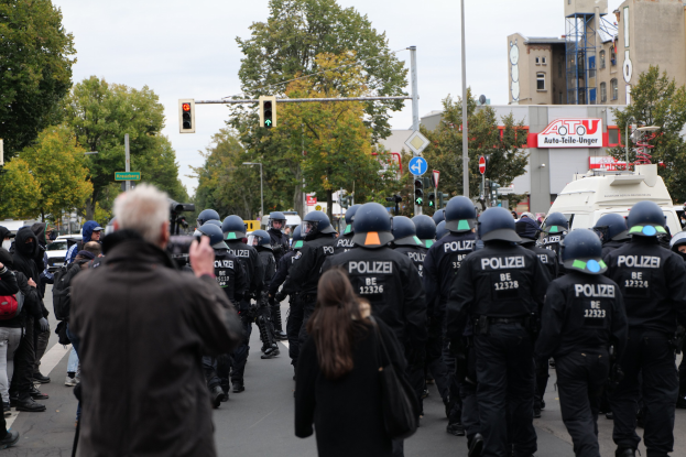 Eine Gruppe von Menschen, die eine Straße entlanggehen, mit Polizeibeamten, einige tragen Helme und haben Taschen oder Kameras, mit Fahrzeugen, Verkehrszeichen, Schildern, Bäumen, Gebäuden und einem klaren Himmel im Hintergrund.