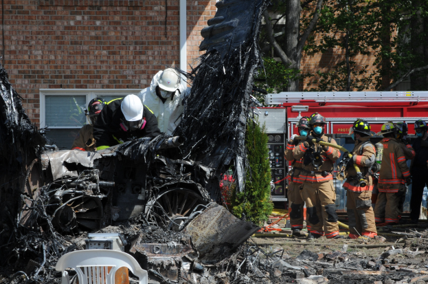 Feuerwehrleute in Schutzausrüstung löschen ein Gebäude Feuer, mit verkohlten Trümmern einschließlich eines Fahrzeugs, eines Stuhls und Pflanzen auf dem Boden.