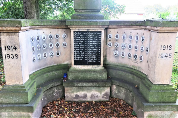 Ein Holocaust-Gedenkmonument in einem jüdischen Friedhof in Berlin, das eine Text- und Nummernwand auf seiner Mauer und umgebende Bäume, einen Zaun und verstreute trockene Blätter zeigt.