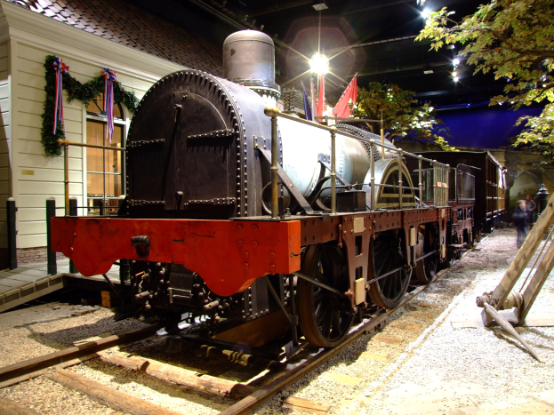 Eine große, alte Dampflokomotive in einem Museum ausgestellt, umgeben von mehreren Menschen, die rechts auf dem Gleis stehen, und von Bäumen und Deckenlampen, die eine festliche Atmosphäre schaffen.