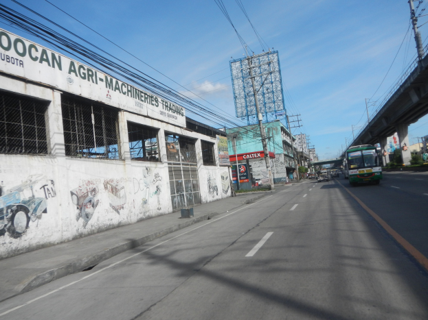 Stadtstraße mit fahrenden Fahrzeugen, eine Brücke auf der rechten Seite, Gebäude, Strommasten mit Drähten, bewölkter Himmel und ein graffiti-beschmiertes Gebäude an der Seite.