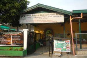 Der Eingang der San Lorenzo Ruiz Pfarrschule zeigt ein Schild mit Text, einen Metallzaun, Menschen unter einem Dach sitzend, Pfosten, ein Schild, Pflanzen, einen Baum und einen bewölkten Himmel.