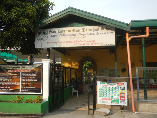 Der Eingang der San Lorenzo Ruiz Pfarrschule zeigt ein Schild mit Text, einen Metallzaun, Menschen unter einem Dach sitzend, Pfosten, ein Schild, Pflanzen, einen Baum und einen bewölkten Himmel.