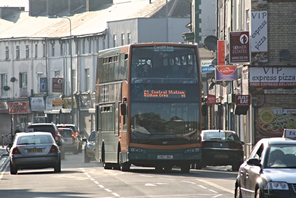 Eine Straße mit Autos und einem Bus im Vordergrund, mit Gebäuden, die Wände, Fenster, Teller und Dächer im Hintergrund sowie Plakate und Banner an den Wänden und einem Pfahl mit einer Straßenlaterne aufweisen.