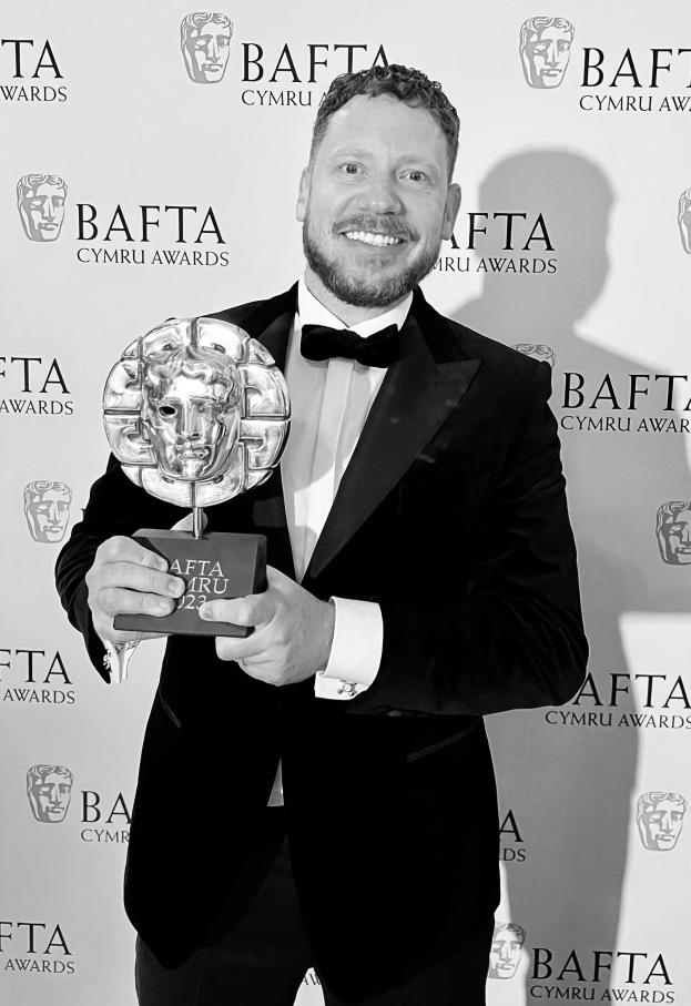 Ein Mann in einem Smoking, der eine BAFTA-Trophäe hält und vor einer Tafel mit 'BAFTA Awards 2018' steht.