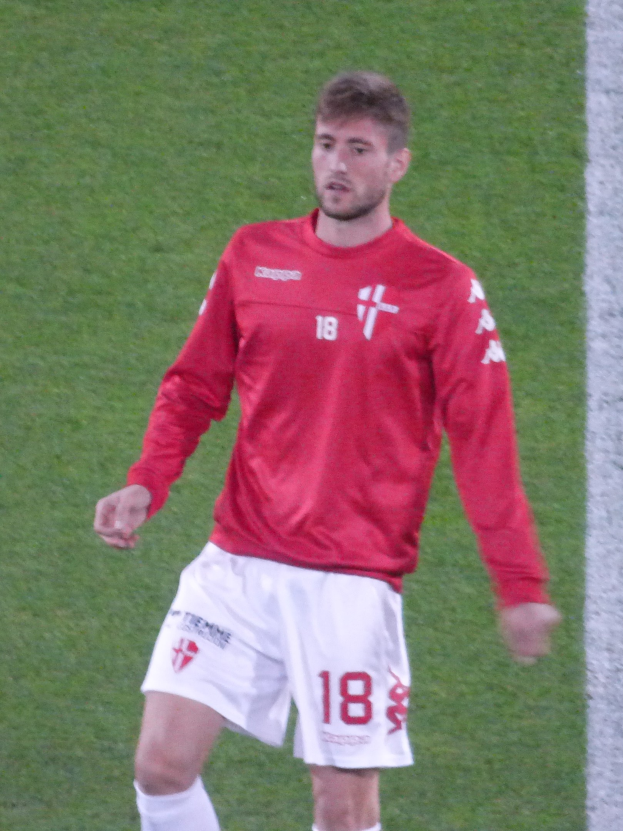 Ein Fußballspieler in rotem Trikot und weißen Shorts steht auf einem Feld mit ausgestreckten Armen und blickt nach vorne.