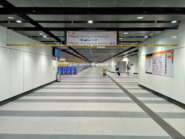 Ein langer Flughafenkorridor mit Menschen, Plakate an der rechten Wand, Deckenleuchten, eine Überwachungskamera und eine Gep├Ąckausgabe in der Mitte.