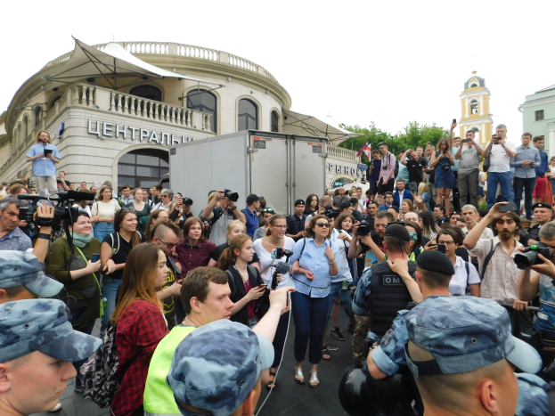 Eine große Gruppe von Menschen steht vor einem Gebäude, einige halten Kameras und Telefone, mit einem Fahrzeug, Bäumen und einem klaren blauen Himmel im Hintergrund, was auf eine Protestaktion gegen das russische Drohnenverbot hindeutet.