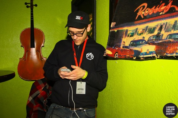 Eine Person mit einer Mütze und einem ID-Karte hält ein Mobiltelefon in der Hand und schaut auf dessen Bildschirm, mit einer Gitarre links und einem Plakat rechts.