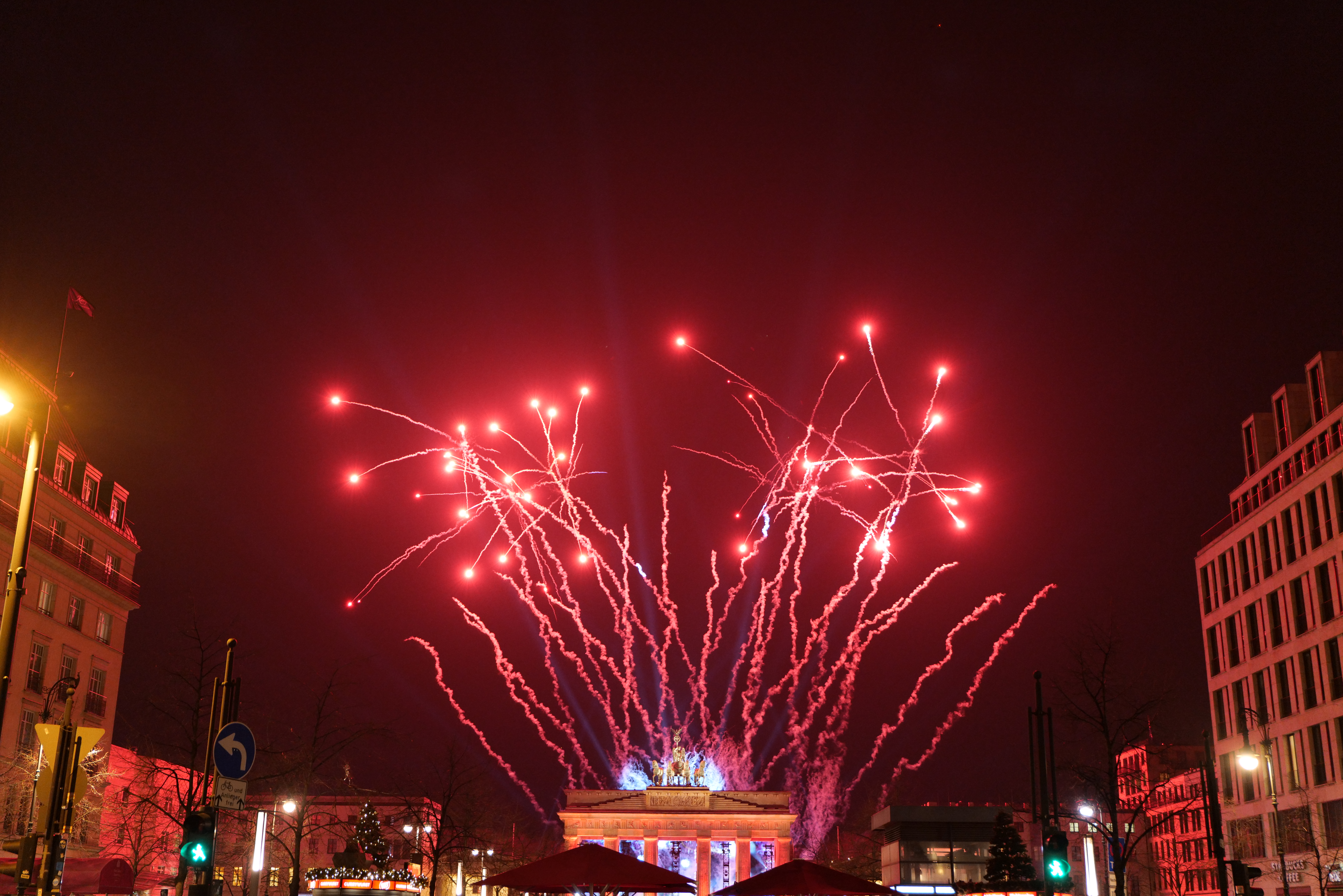 Eine Stadtstraße in Berlin an Silvester, voller Gebäude, Bäume, Laternenpfähle, Verkehrszeichen, Zelte und Menschen, mit einem Feuerwerk erhellten Himmel im Hintergrund.