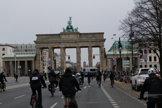 Eine Gruppe von Menschen, die auf Fahrrädern eine Straße vor dem Brandenburger Tor in Berlin, Deutschland, entlangfahren, mit Bäumen, Laternen, Verkehrsschildern, Schildern und Gebäuden mit Fenstern im Hintergrund und dem Himmel in der Ferne.