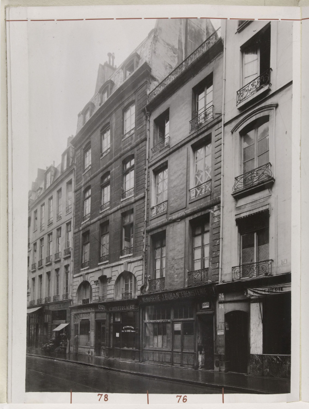 Schwarz-weißes Foto einer Stadtstraße mit Gebäuden mit Fenstern, Geländern und Namensschildern sowie einer Straße unten.
