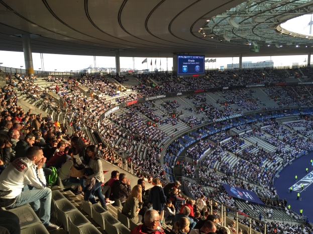 Eine große Menschenmenge sitzt in einem Stadion und schaut ein Fußballspiel, mit einer Bühne auf der rechten Seite, Fahnen, Stangen und einem Bildschirm im Hintergrund, unter einem sichtbaren Himmel, im Allianz Stadion in München, Deutschland.